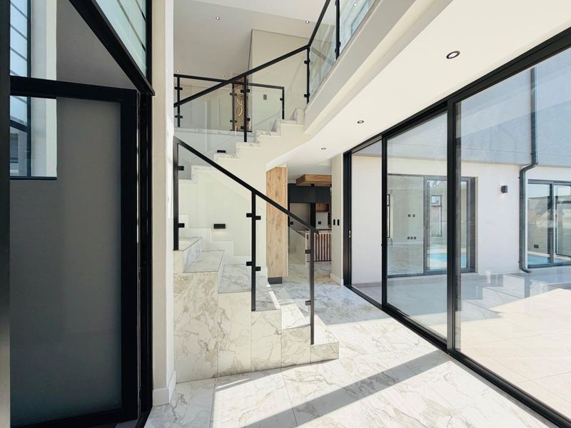 Modern double-volume entrance hall featuring marble-look tiled flooring, a sleek glass staircase balustrade, and expansive sliding doors leading to the outdoor area.