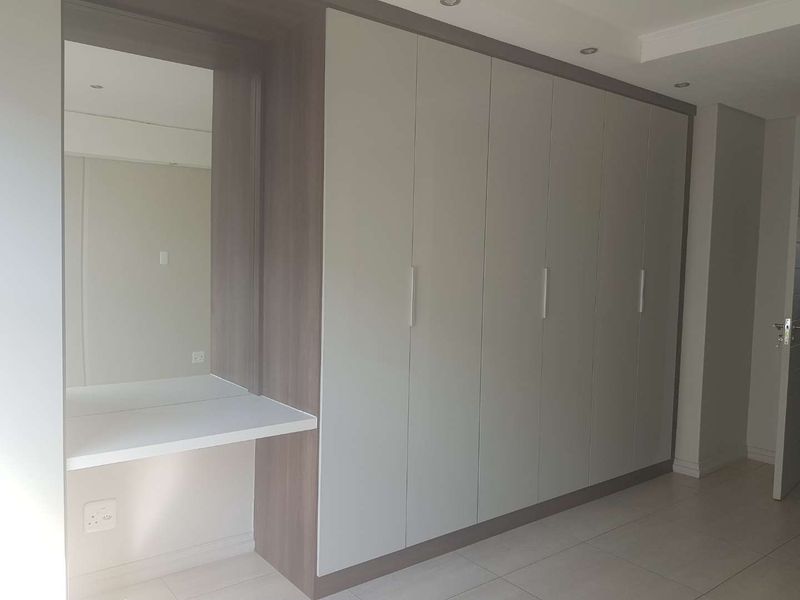 A modern bedroom featuring extensive built-in cupboards with a integrated vanity station, recessed lighting, and neutral tiled flooring.