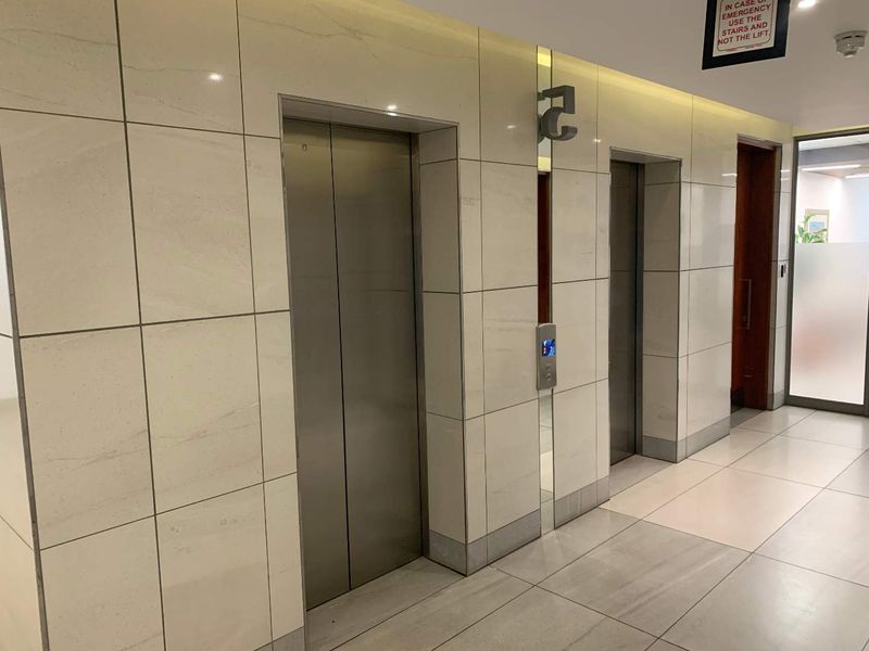 A modern office building elevator lobby featuring stainless steel lift doors, polished tiled flooring, and contemporary wall tiling.