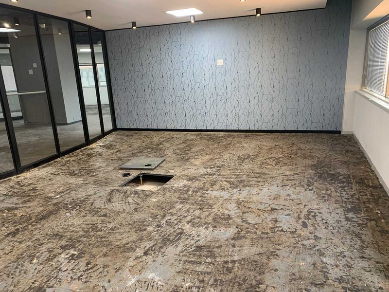 An empty office space featuring a glass partition wall, recessed ceiling lighting, and a subfloor requiring new flooring installation.