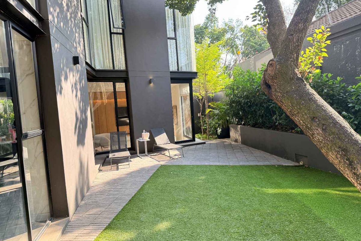 A modern rear garden featuring a paved patio area, artificial lawn, and large floor-to-ceiling glass doors connecting to the interior living space.