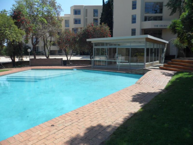 Communal swimming pool area featuring a glass-enclosed pavilion, brick paved surround, and landscaped garden within an apartment complex setting.