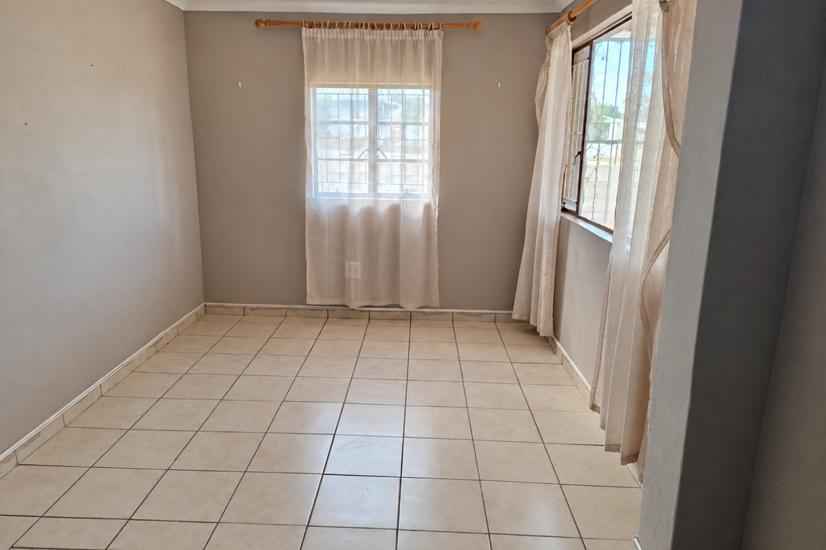 A vacant bedroom featuring neutral wall tones, tiled flooring, and windows fitted with security bars and curtain rods.