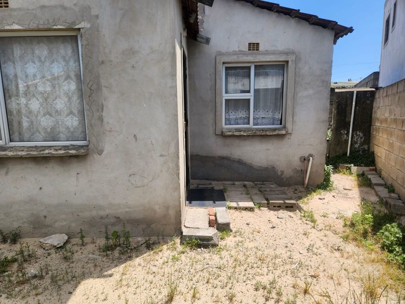 Rear exterior view of the property featuring unfinished plaster walls, aluminium window frames, and a concrete block boundary wall in a basic yard setting.