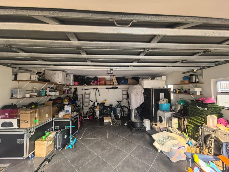 A spacious double garage featuring tiled flooring, overhead sectional door mechanism, and extensive wall-mounted shelving for storage.