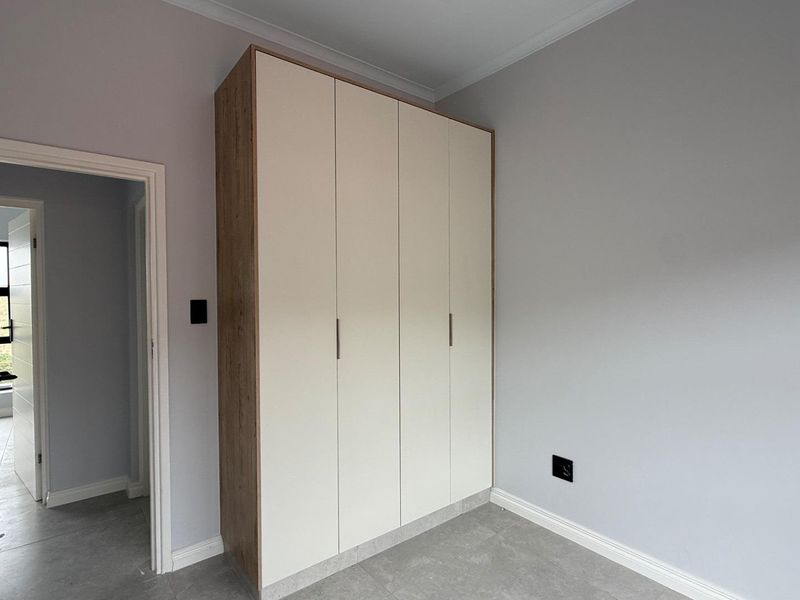 A clean, modern bedroom featuring neutral grey walls, tiled flooring, and a large, contemporary built-in wardrobe with light-toned doors.