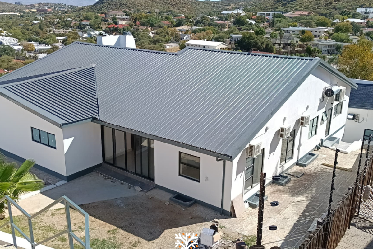 Aerial view of a modern house with a corrugated metal roof, featuring large glass sliding doors and multiple wall-mounted air conditioning units.