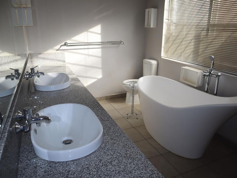 Modern bathroom featuring a double vanity with granite countertop, a sleek freestanding bathtub, and neutral tiled flooring with natural light from venetian blinds.