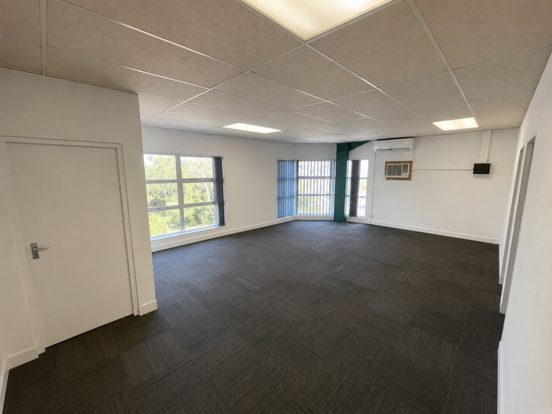 A spacious, empty office area featuring carpet tiles, a suspended ceiling with recessed lighting, and a wall-mounted split-system air conditioner.