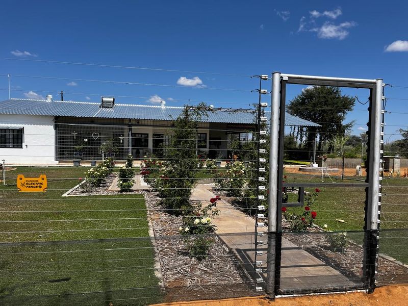 Front view of the farm property featuring a secure electric fence, metal gate, paved garden path, and a building with a corrugated metal roof.