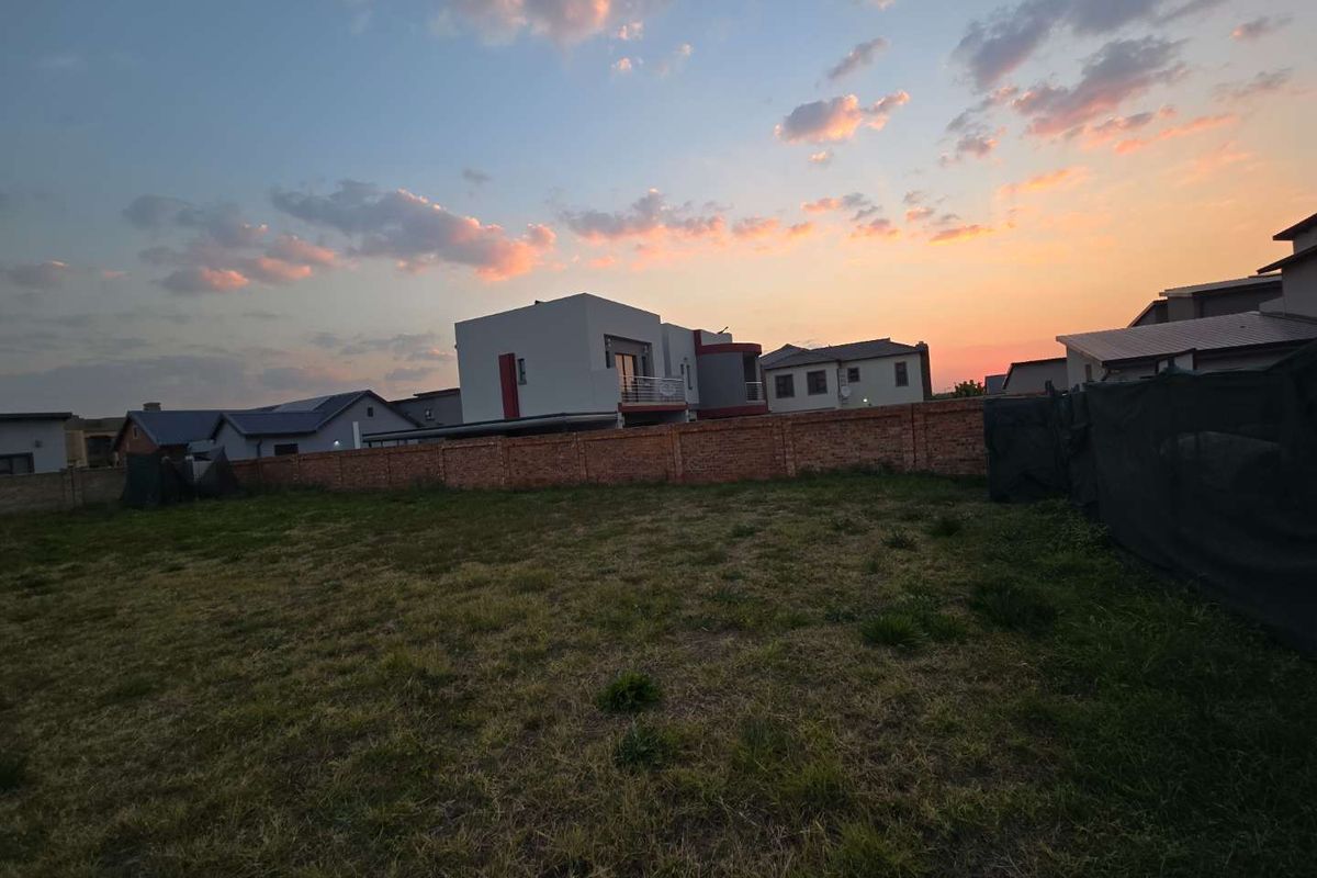 A vacant residential plot featuring a grass lawn and a brick boundary wall, set against a sunset sky in a developed neighborhood.