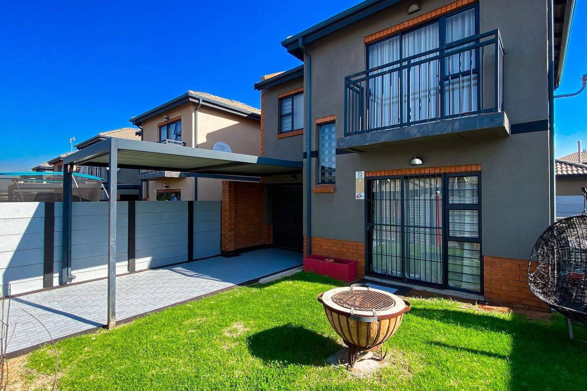 Rear view of the property featuring a paved carport, a small lawn, and an upper-level balcony with modern brick and render finishes.