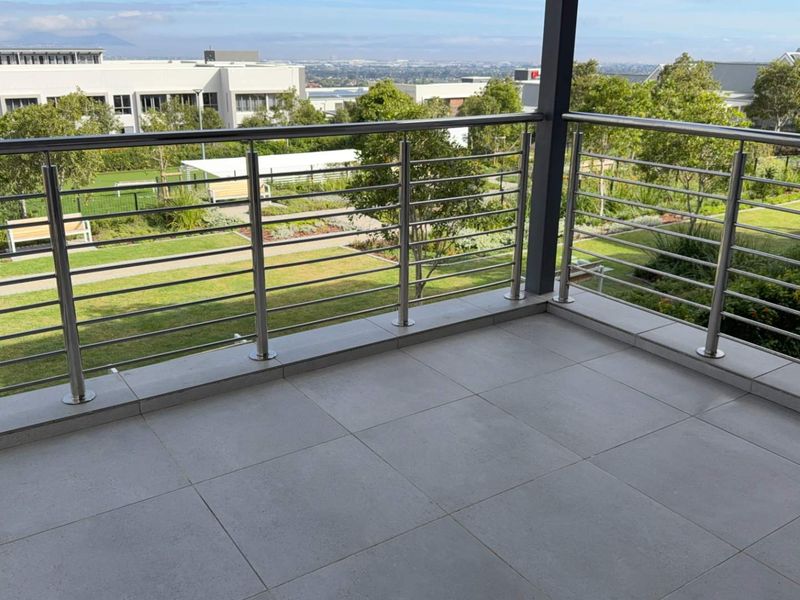 A spacious covered balcony featuring modern stainless steel balustrades and neutral floor tiles, offering pleasant views over the retirement village grounds.