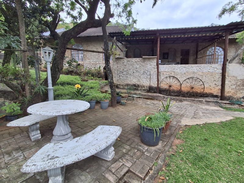 A rustic stone-clad farmhouse exterior featuring a covered patio area, paved courtyard, and arched windows, set within a mature garden environment.
