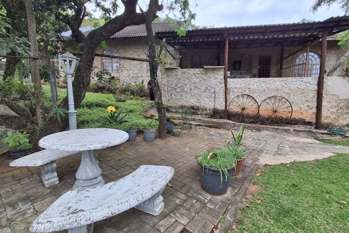 A rustic stone-clad farmhouse exterior featuring a covered patio area, paved courtyard, and arched windows, set within a mature garden environment.