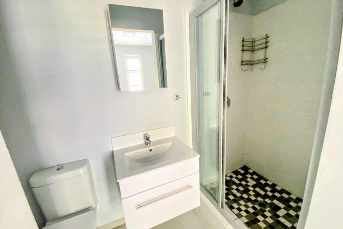A clean, modern bathroom featuring a white vanity, wall-mounted mirror, toilet, and a glass-enclosed shower with a distinctive checkered tile floor.