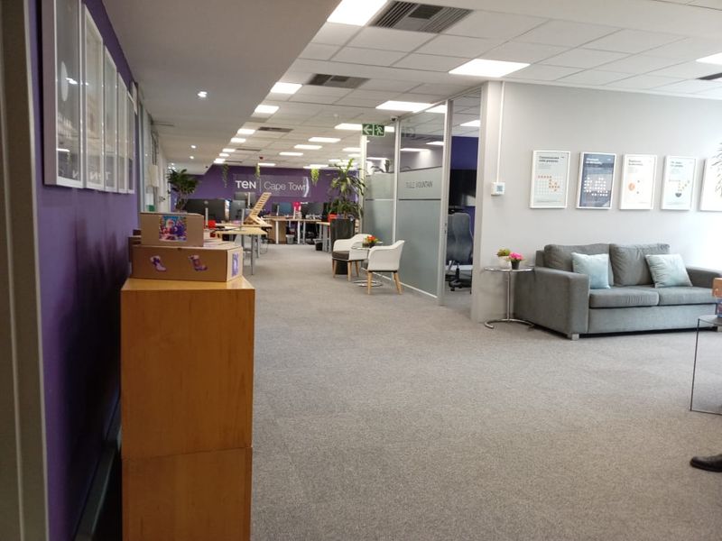 A modern open-plan office space featuring glass partitions, suspended ceilings with recessed lighting, and carpeted flooring throughout the workspace.