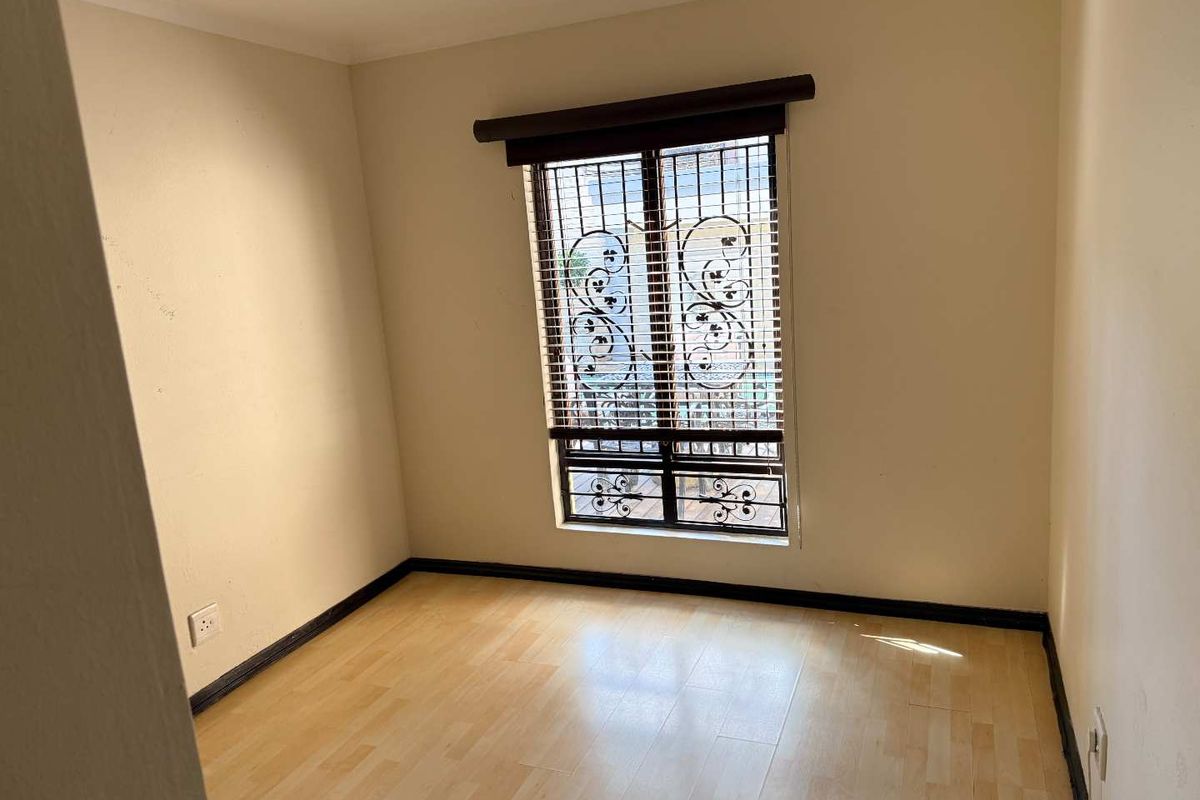 A vacant bedroom featuring light-toned laminate flooring, neutral walls, and a window fitted with security bars and blinds.