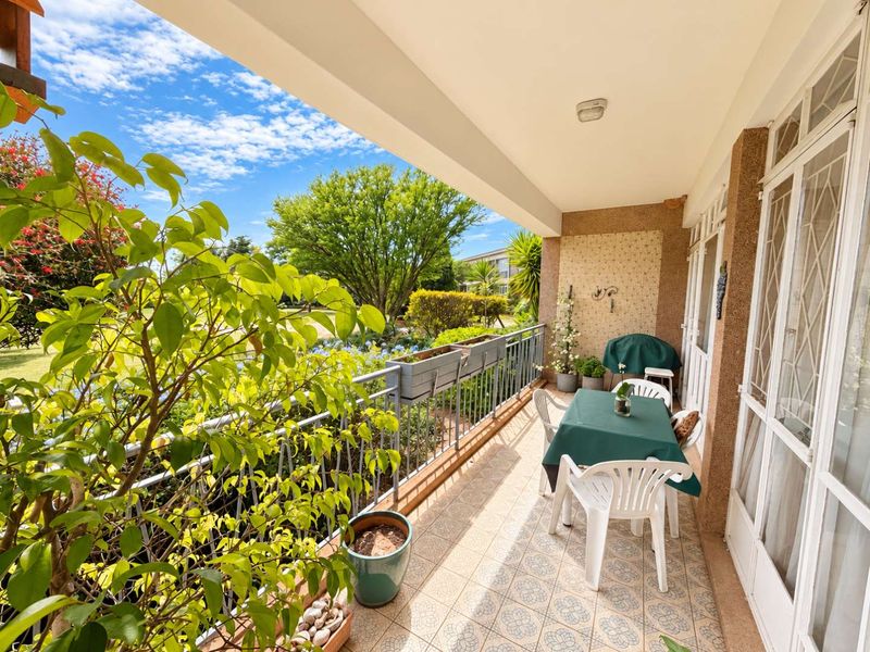 A covered balcony featuring tiled flooring, a metal balustrade, and direct access to the apartment through sliding glass doors, overlooking a landscaped garden area.
