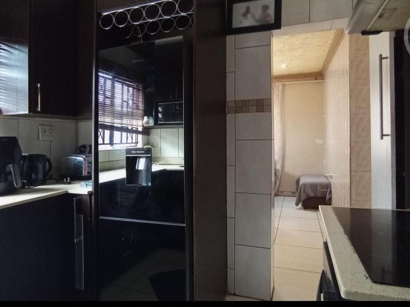 A functional kitchen featuring dark cabinetry, light countertops, an electric hob, and tiled finishes, with an adjacent room visible through the doorway.