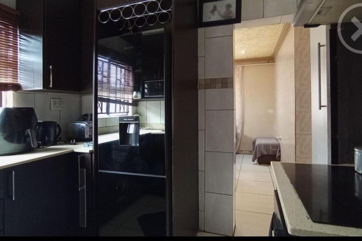 A functional kitchen featuring dark cabinetry, light countertops, an electric hob, and tiled finishes, with an adjacent room visible through the doorway.