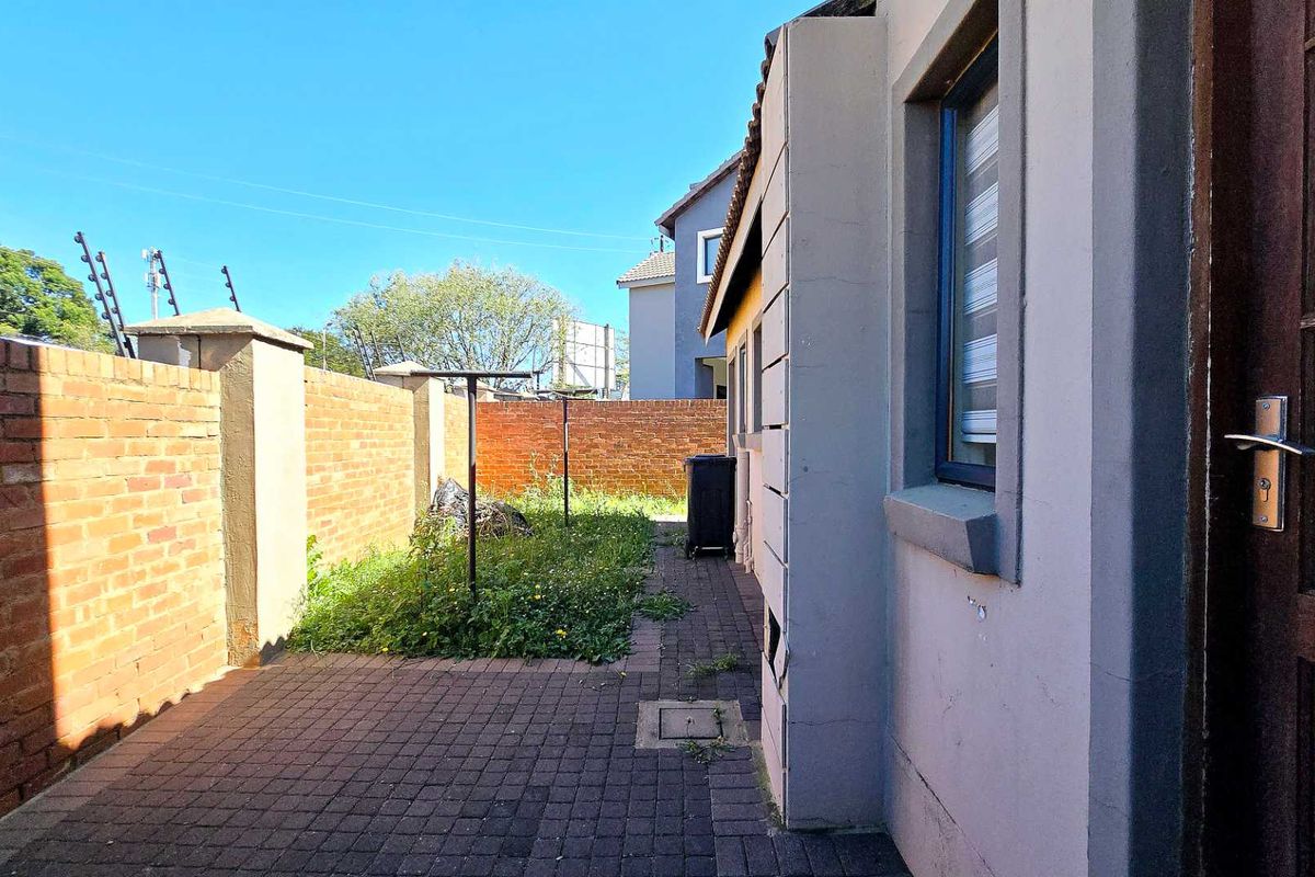 A narrow rear exterior area featuring paved flooring, a brick boundary wall with electric fencing, and an overgrown garden section.
