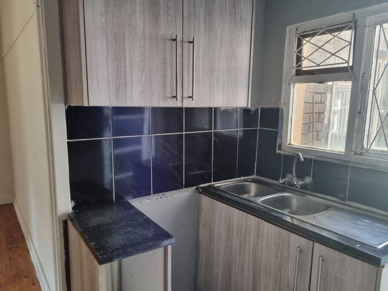 A compact kitchen featuring light wood-look cabinetry, a double stainless steel sink, and a dark tiled backsplash, currently requiring minor finishing and maintenance.