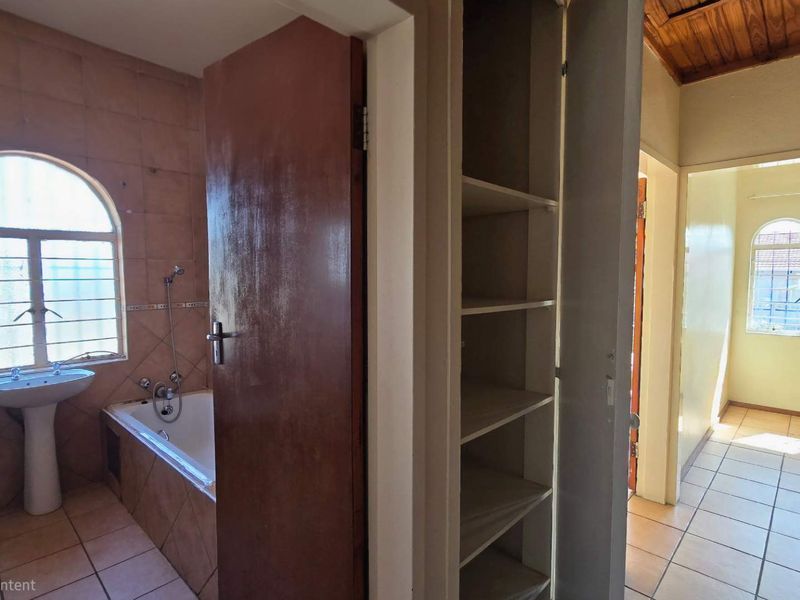 A functional bathroom featuring a pedestal basin, built-in bathtub, and tiled finishes, adjacent to a hallway with built-in storage shelving.