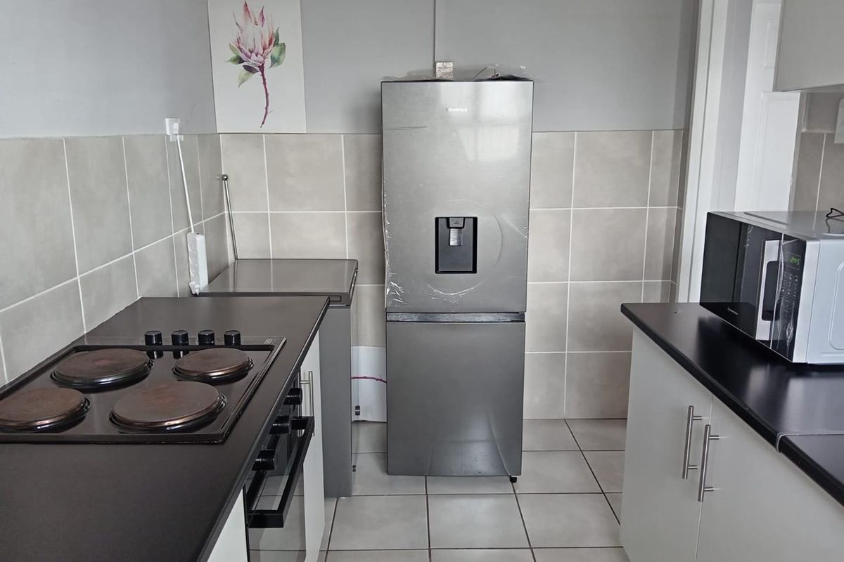 A functional kitchen featuring built-in cabinetry, dark laminate countertops, an electric hob, oven, and neutral tiled splashbacks and flooring.