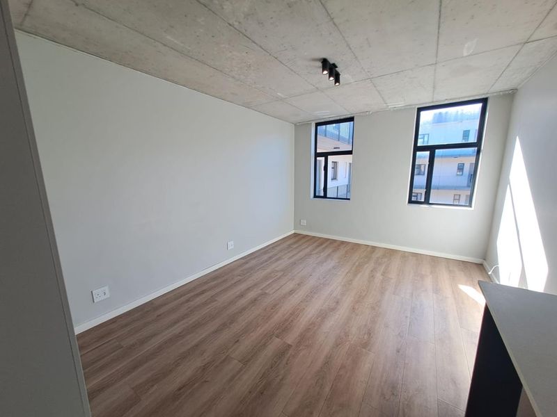 A bright, modern bedroom featuring an exposed concrete ceiling, laminate flooring, and large black-framed windows that provide ample natural light.
