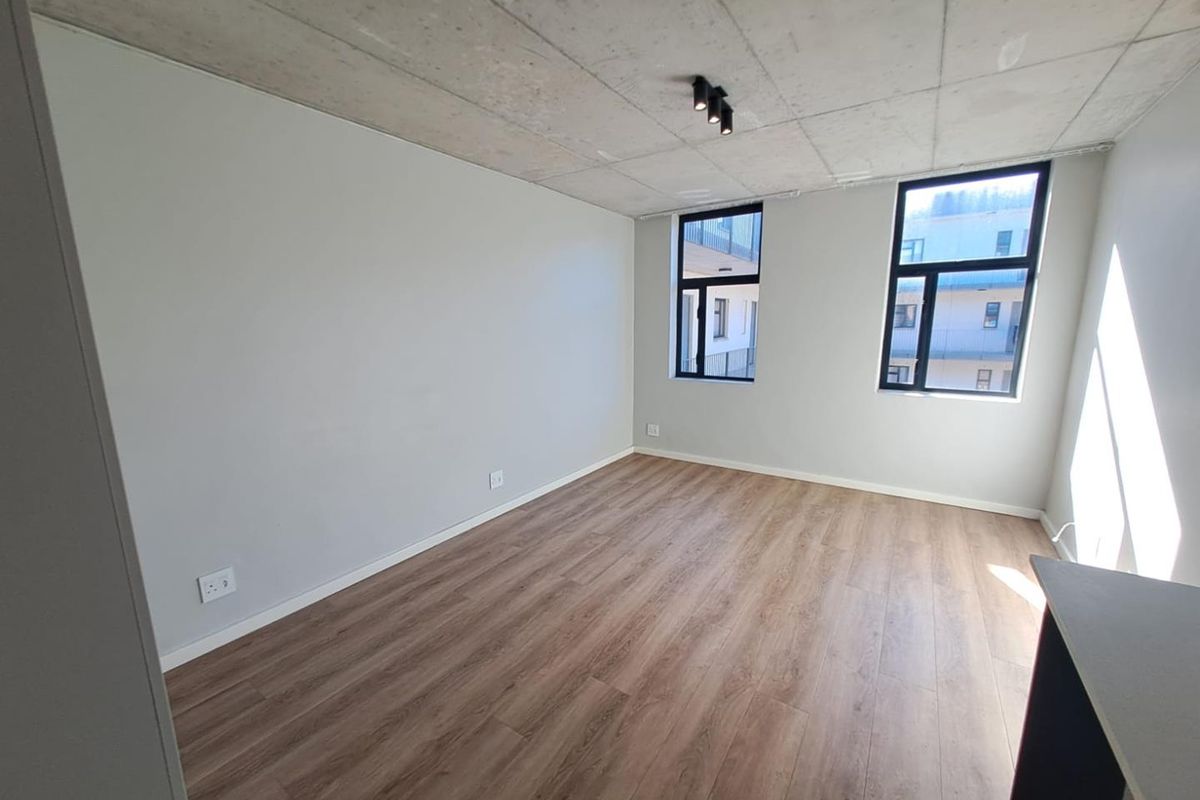 A bright, modern bedroom featuring an exposed concrete ceiling, laminate flooring, and large black-framed windows that provide ample natural light.