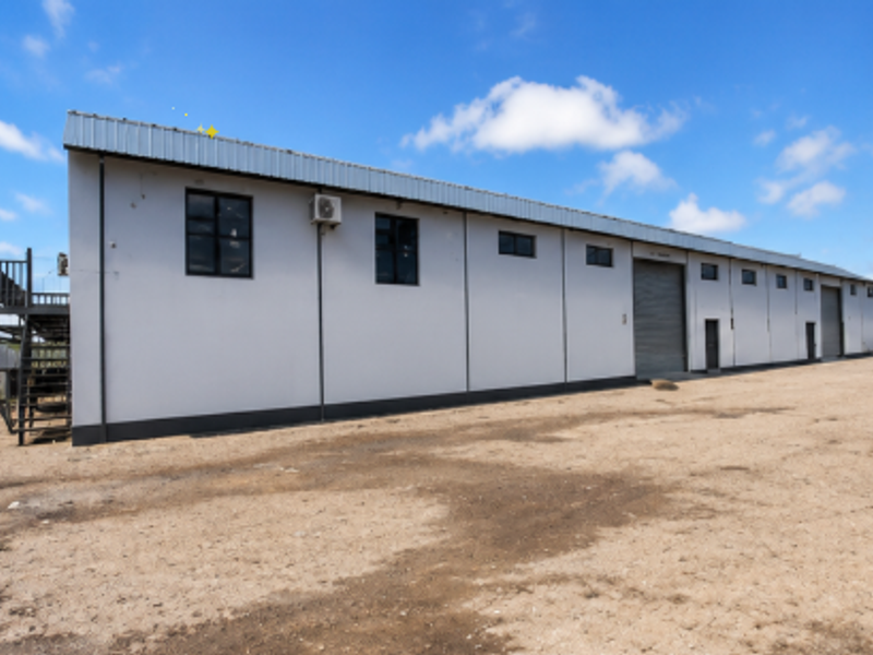Rear view of an industrial warehouse building featuring multiple roller shutter doors, an external staircase, and a large unpaved yard area.