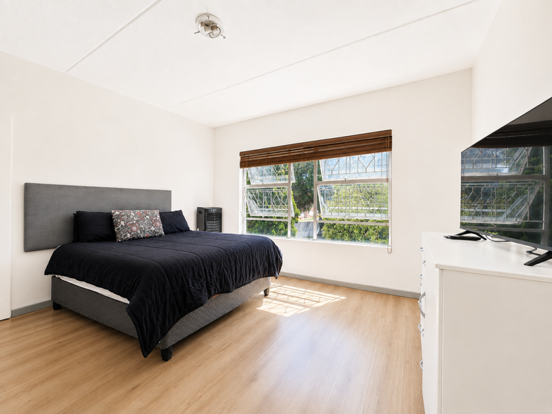A well-lit bedroom featuring light wood-look laminate flooring, a large window with security bars, and wooden venetian blinds.