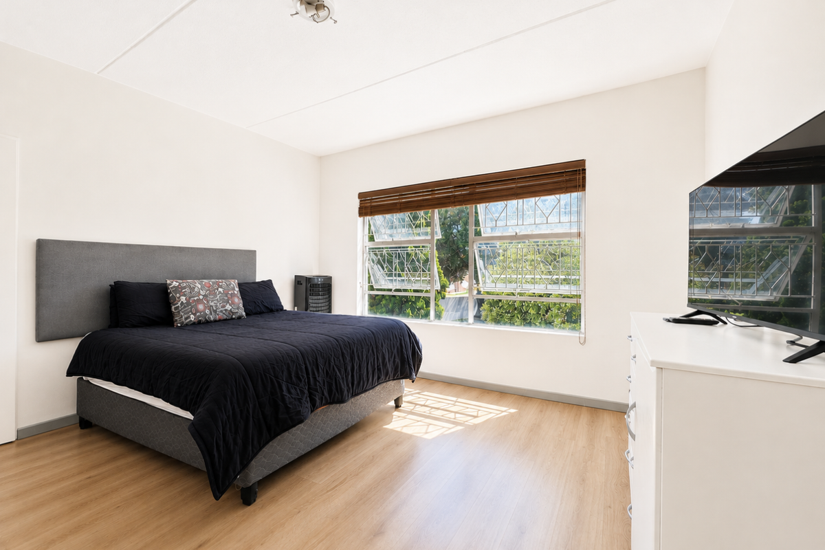 A well-lit bedroom featuring light wood-look laminate flooring, a large window with security bars, and wooden venetian blinds.