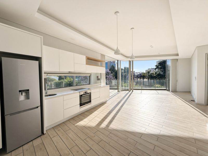 Modern open-plan kitchen and living area featuring sleek white cabinetry, integrated appliances, wood-look tile flooring, and expansive floor-to-ceiling windows with balcony access.