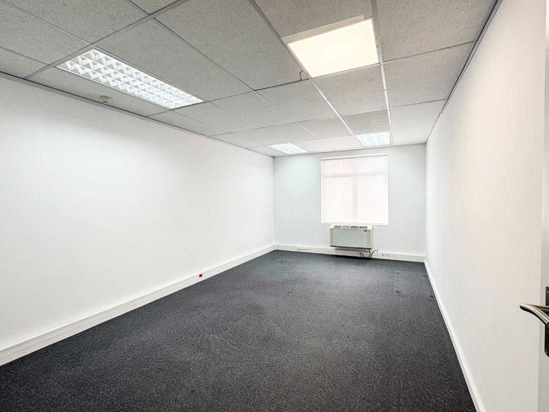 A clean, empty office space featuring a suspended ceiling with recessed lighting, dark carpet flooring, and a wall-mounted air conditioning unit.