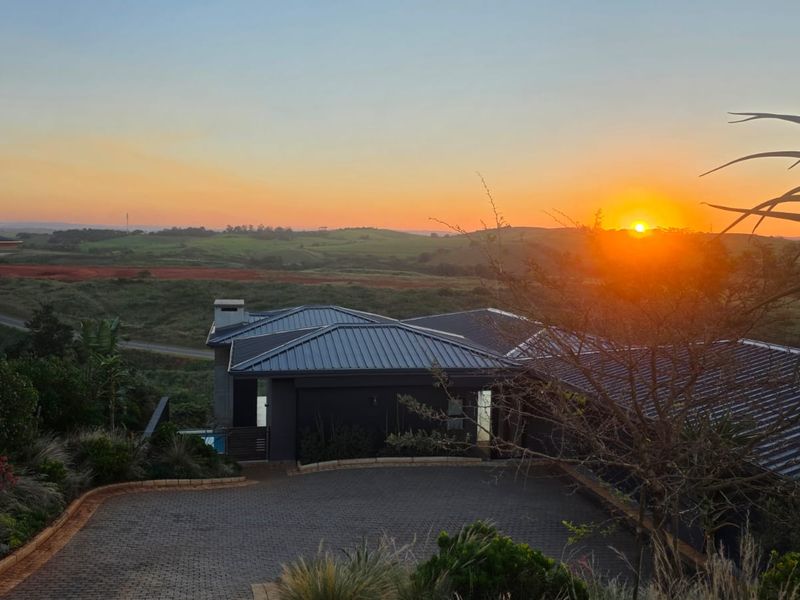 An elevated view of the property featuring a modern metal roof, paved driveway, and chimney, set against a scenic sunset landscape.