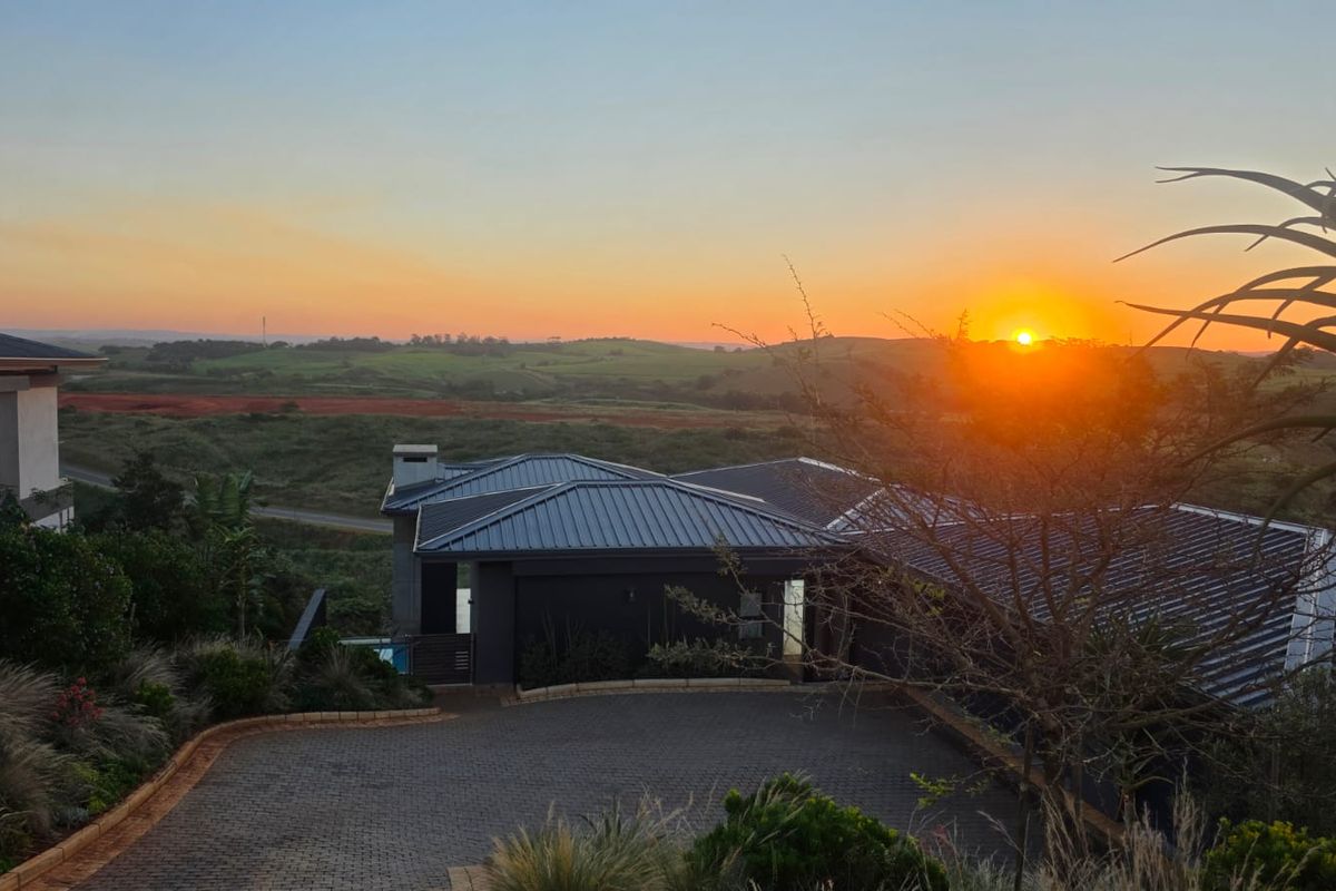An elevated view of the property featuring a modern metal roof, paved driveway, and chimney, set against a scenic sunset landscape.