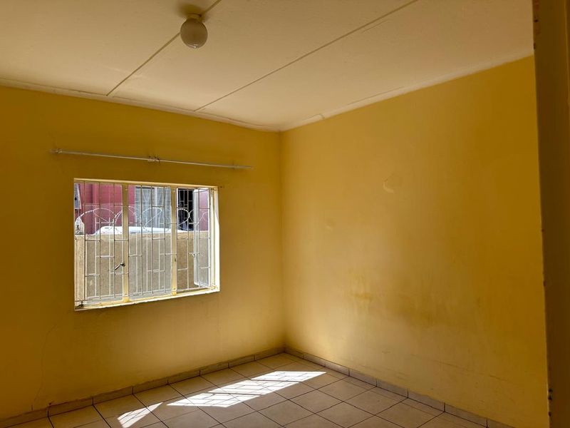 An empty bedroom featuring tiled flooring, yellow painted walls with visible wear, and a window fitted with security bars.