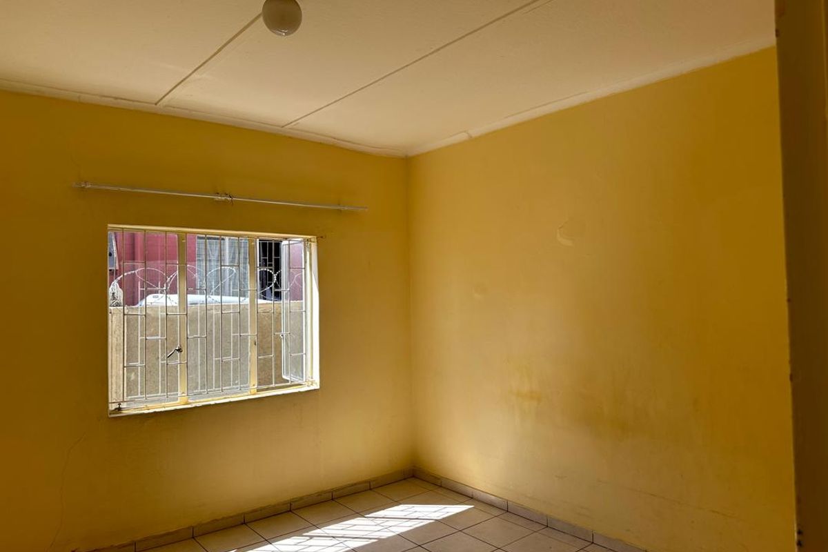 An empty bedroom featuring tiled flooring, yellow painted walls with visible wear, and a window fitted with security bars.