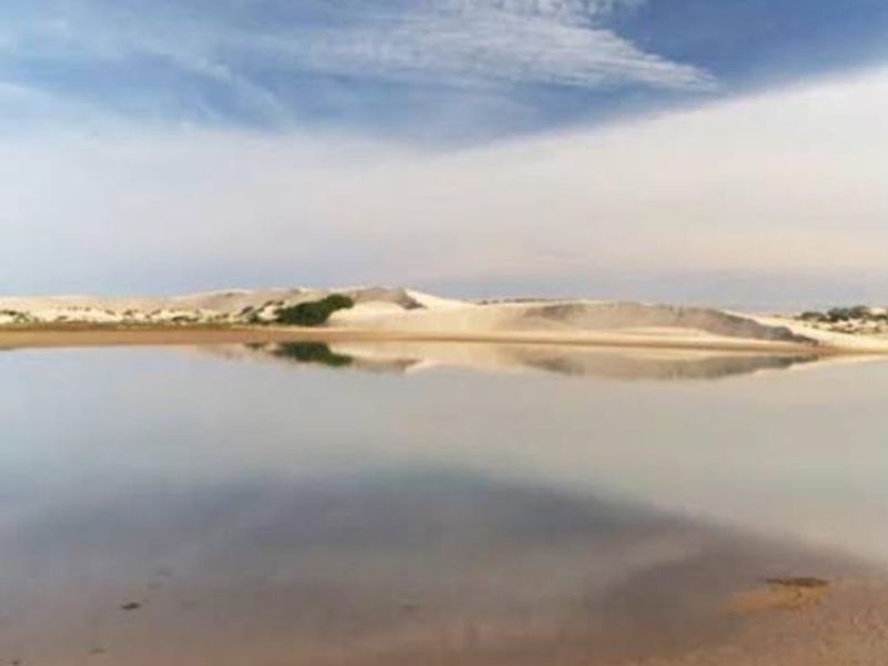 A scenic view of a tranquil coastal lagoon bordered by natural sand dunes under a clear sky.