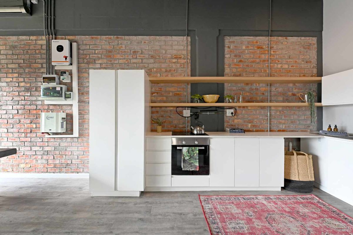 Modern industrial-style kitchen featuring exposed brick walls, sleek white cabinetry, built-in oven, electric hob, and open shelving with an integrated inverter system.