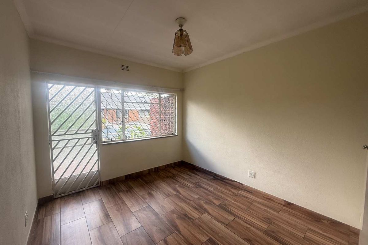 A bright, empty bedroom featuring modern laminate flooring, a security gate, and burglar bars on the window, providing a clean canvas for personalization.