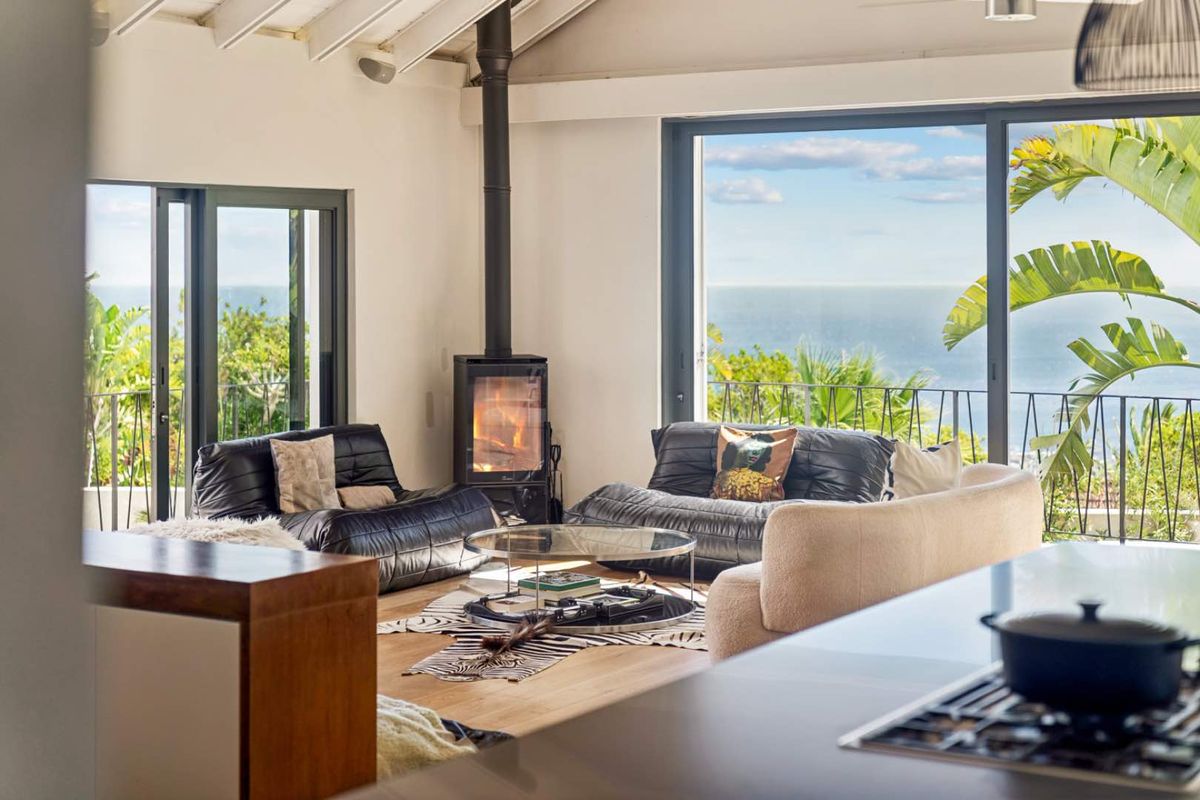 Bright living room featuring a modern freestanding fireplace, exposed beam ceiling, and large sliding glass doors opening to a scenic ocean view balcony.