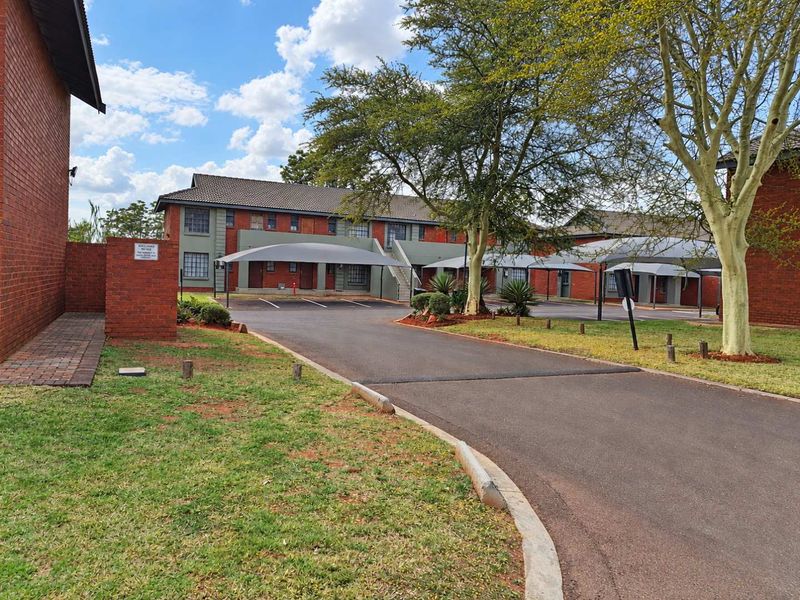 Exterior view of an apartment complex featuring brick buildings, paved driveway, and covered parking bays set within a landscaped residential estate.