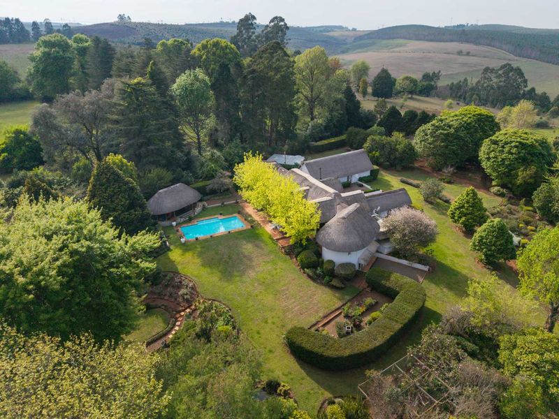 Aerial view of a farm property featuring multiple thatched-roof buildings, a swimming pool, a thatched lapa, and extensive landscaped gardens set in a rural landscape.