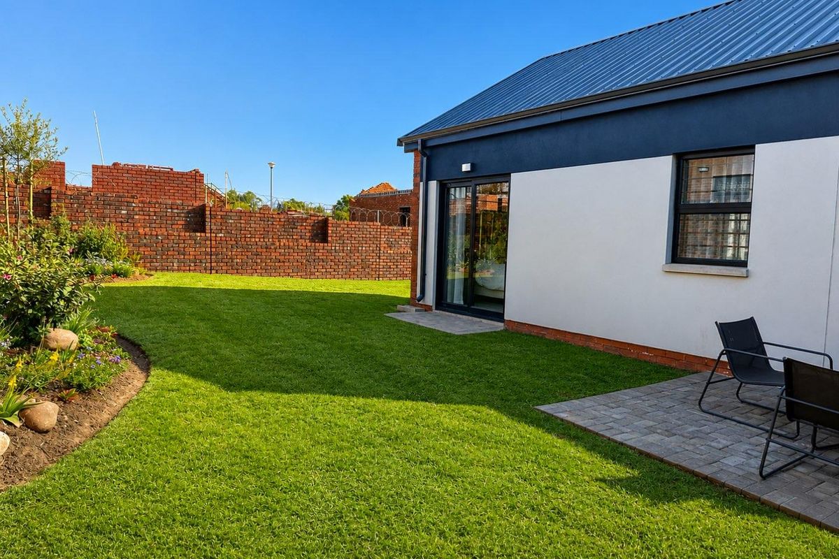 A well-maintained rear garden featuring a manicured lawn, paved patio area, and perimeter brick walling with security fencing.