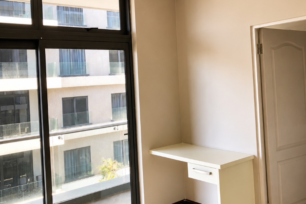 A bright, empty bedroom featuring tiled flooring, crown molding, a built-in desk, and a large sliding glass door leading to an exterior balcony.