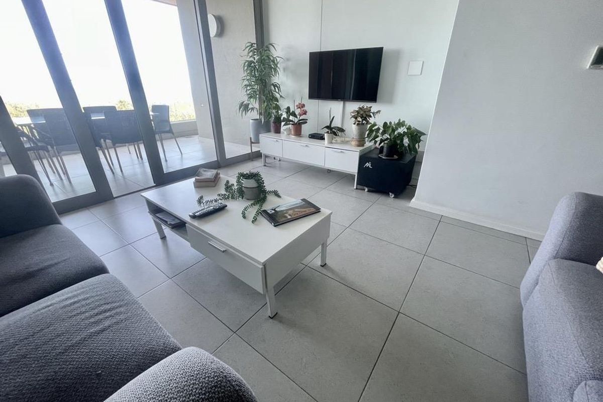 A bright, modern living room featuring large sliding glass doors that open onto a spacious balcony, finished with neutral floor tiles.