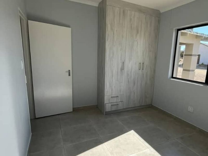 A bright, empty bedroom featuring neutral tiled flooring, a modern built-in wardrobe with wood-grain finish, and clean, light-grey painted walls.